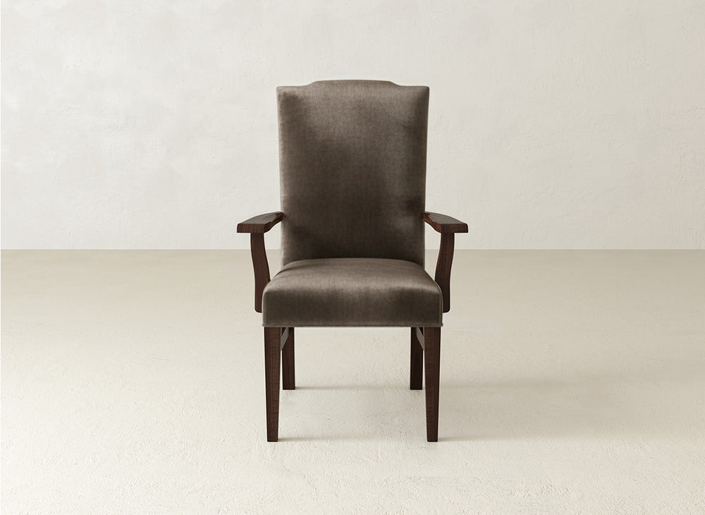 The Hadley Arm Dining Chair by James+James features dark brown upholstery, a high back, padded seat and backrest, and wooden armrests, standing in an empty, light-colored room with a plain wall and floor.