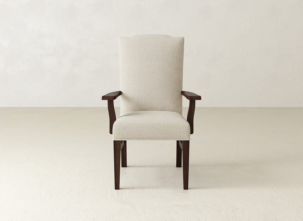 The Hadley Arm Dining Chair by James+James, featuring beige upholstery with dark wooden legs and armrests, sits centered in a light-toned room with textured walls and flooring.
