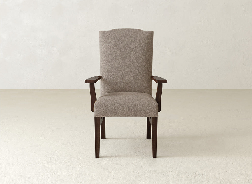 The Hadley Arm Dining Chair by James+James features beige upholstery, dark wood legs, and armrests, offering comfort and style. Shown on a light textured floor against an off-white wall.
