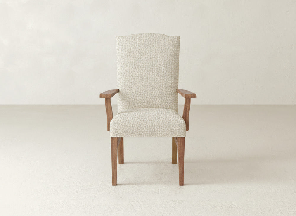 The Hadley Arm Dining Chair by James+James, featuring beige upholstery and wooden legs and armrests, is centered in a minimalist room with light walls and flooring.