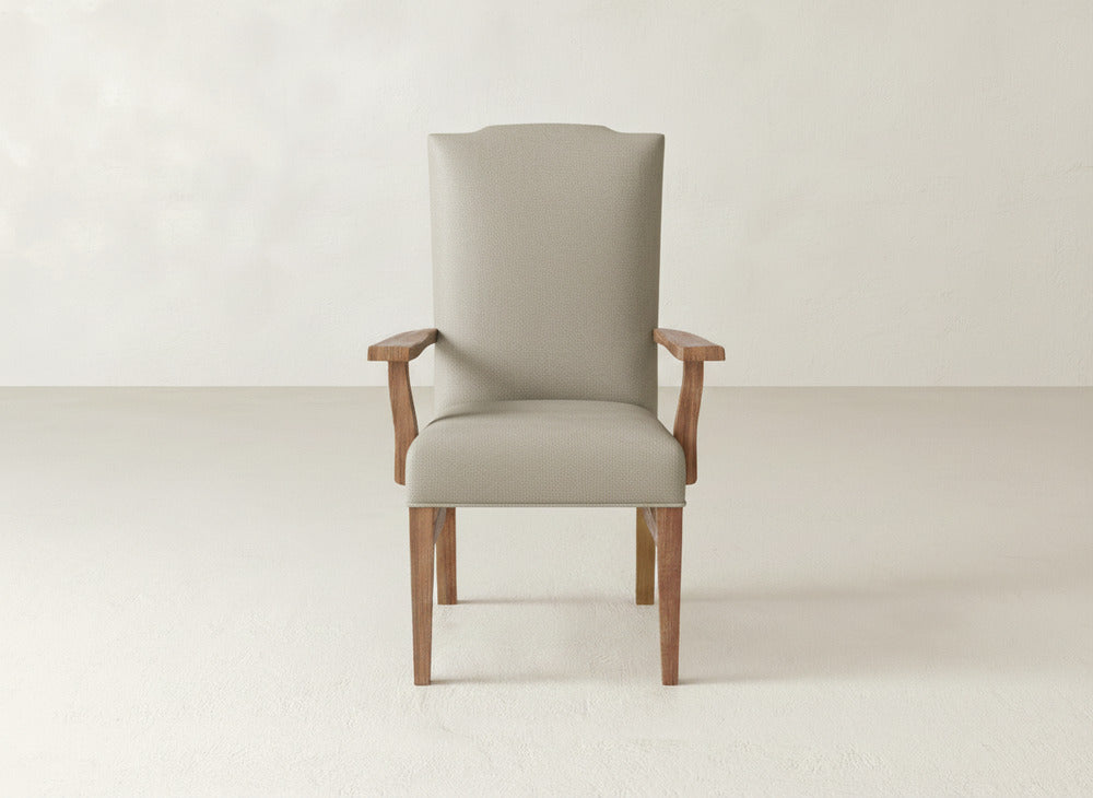 The Hadley Arm Dining Chair by James+James, featuring beige upholstery and wooden legs and armrests, sits against a simple, light wall and floor.
