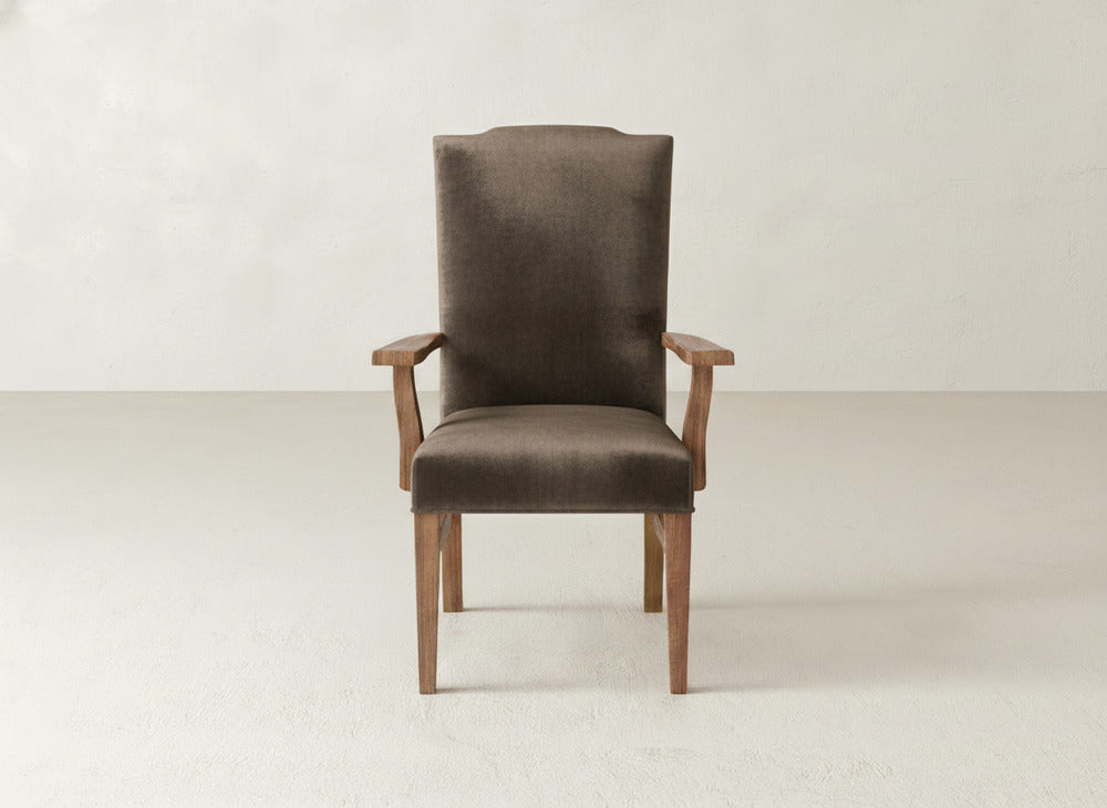The Hadley Arm Dining Chair by James+James features brown upholstery, wooden legs, and armrests, placed against a plain light-colored wall and floor.