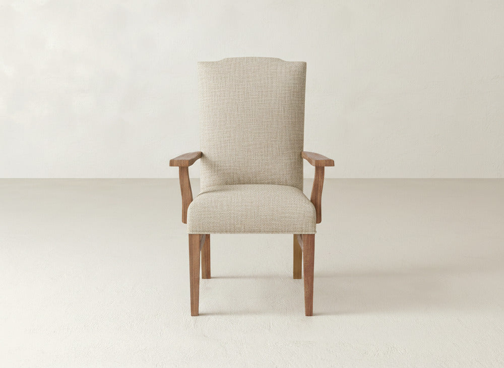A James+James Hadley Arm Dining Chair, upholstered in beige with wooden legs and armrests, is centered against a plain, light-colored wall and floor background.