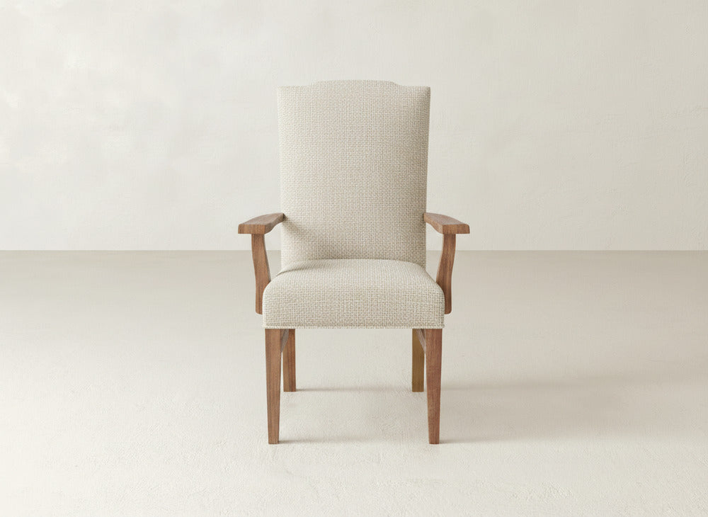 The Hadley Arm Dining Chair by James+James, featuring beige upholstery, wooden legs, and armrests, sits against a plain light wall on a smooth neutral floor.