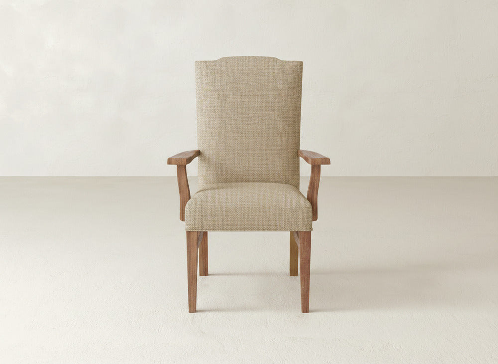 A beige James+James Hadley Arm Dining Chair with wooden legs and armrests is centered in a minimal, light-colored room with a plain wall and floor.