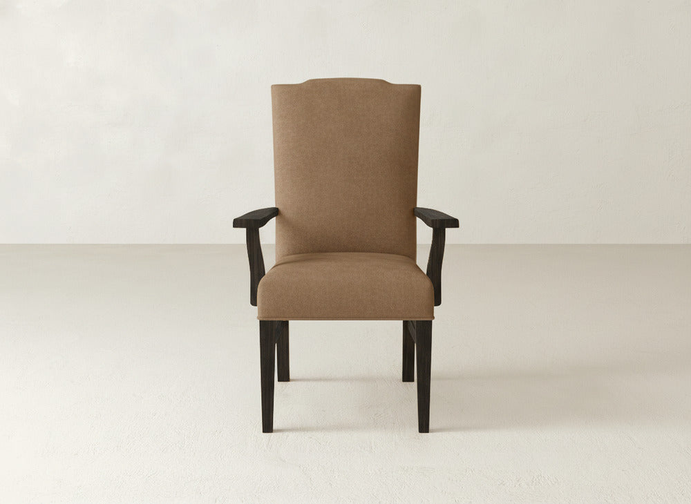 The Hadley Arm Dining Chair by James+James features brown upholstery, a tall backrest, and dark wooden arms and legs, shown on a light floor against a plain off-white wall.