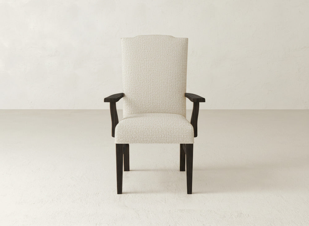 The Hadley Arm Dining Chair by James+James, featuring beige upholstery and dark wooden legs and armrests, is centered against a plain, light-colored wall and floor.