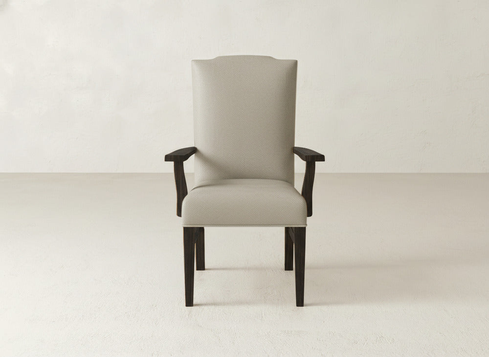 The Hadley Arm Dining Chair by James+James features beige upholstery, dark wood legs, and armrests, offering the look of a comfortable armchair for any space. It is pictured facing forward against a plain light wall and floor.