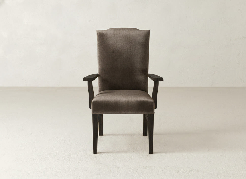 The Hadley Arm Dining Chair by James+James features brown upholstery, a high back, and dark wooden legs. It is shown on a light, neutral floor against a plain off-white wall.