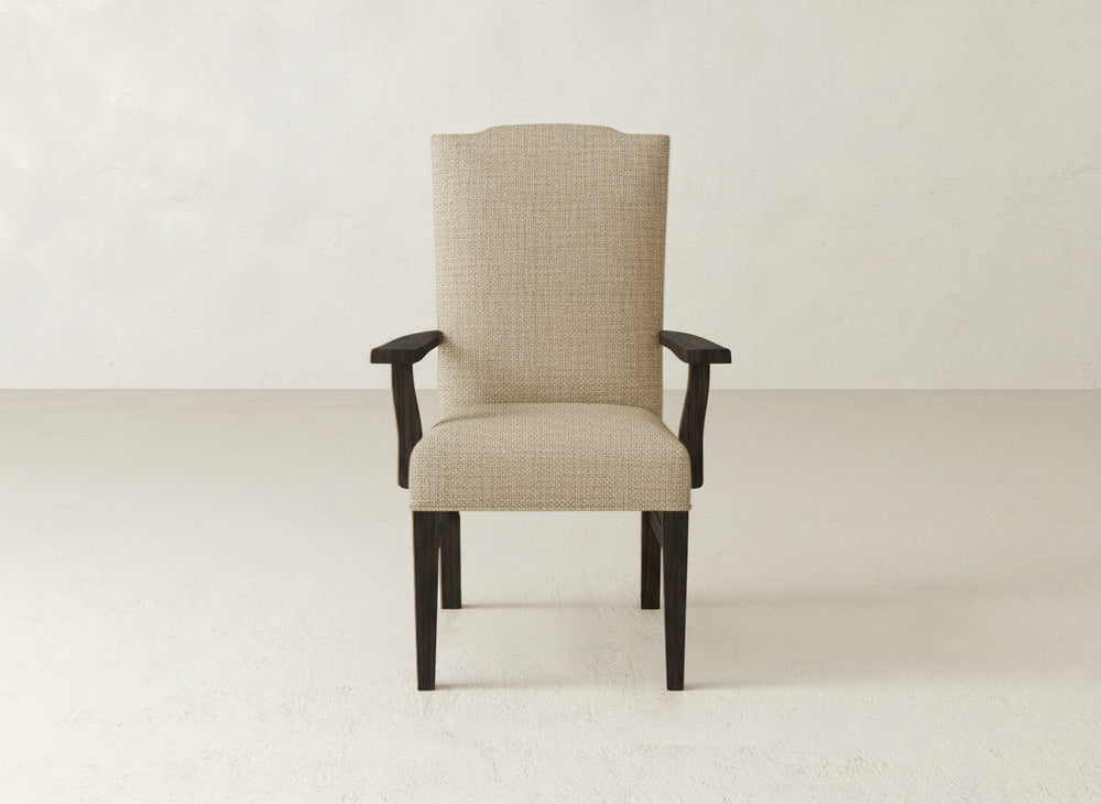 The Hadley Arm Dining Chair by James+James features beige upholstery, dark wooden legs and armrests, and a tall, slightly curved backrest for comfort. It's shown against a plain, light-colored wall and floor.