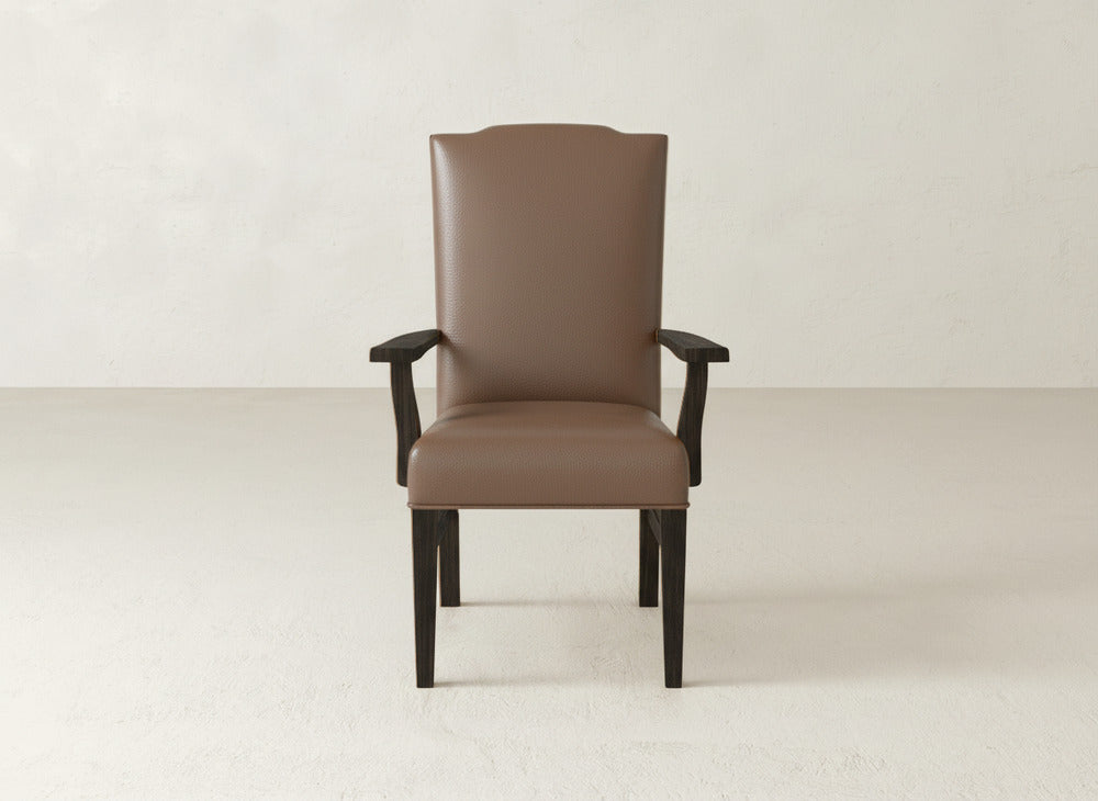 The Hadley Arm Dining Chair by James+James, featuring brown upholstery and dark wooden legs and armrests, is set against a simple, light-colored wall and floor backdrop.