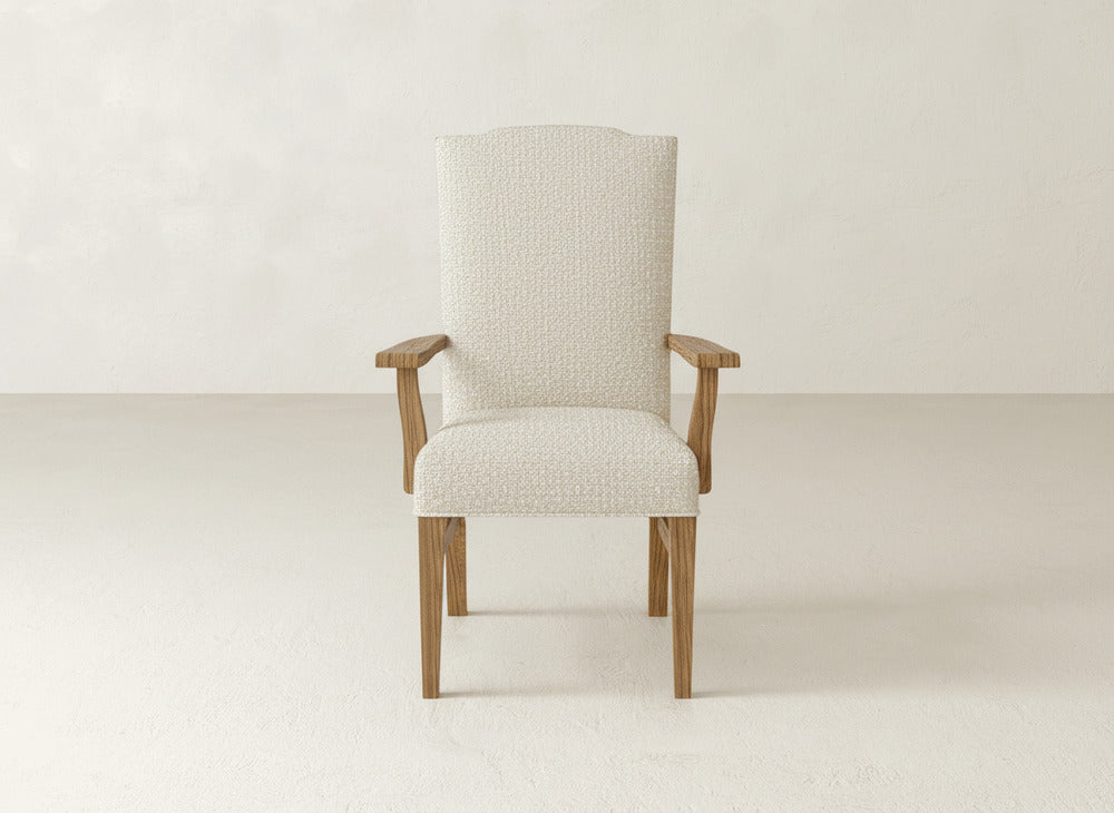 The Hadley Arm Dining Chair by James+James features a textured cream upholstered seat and back with light wood armrests and legs, set against an off-white wall and floor.