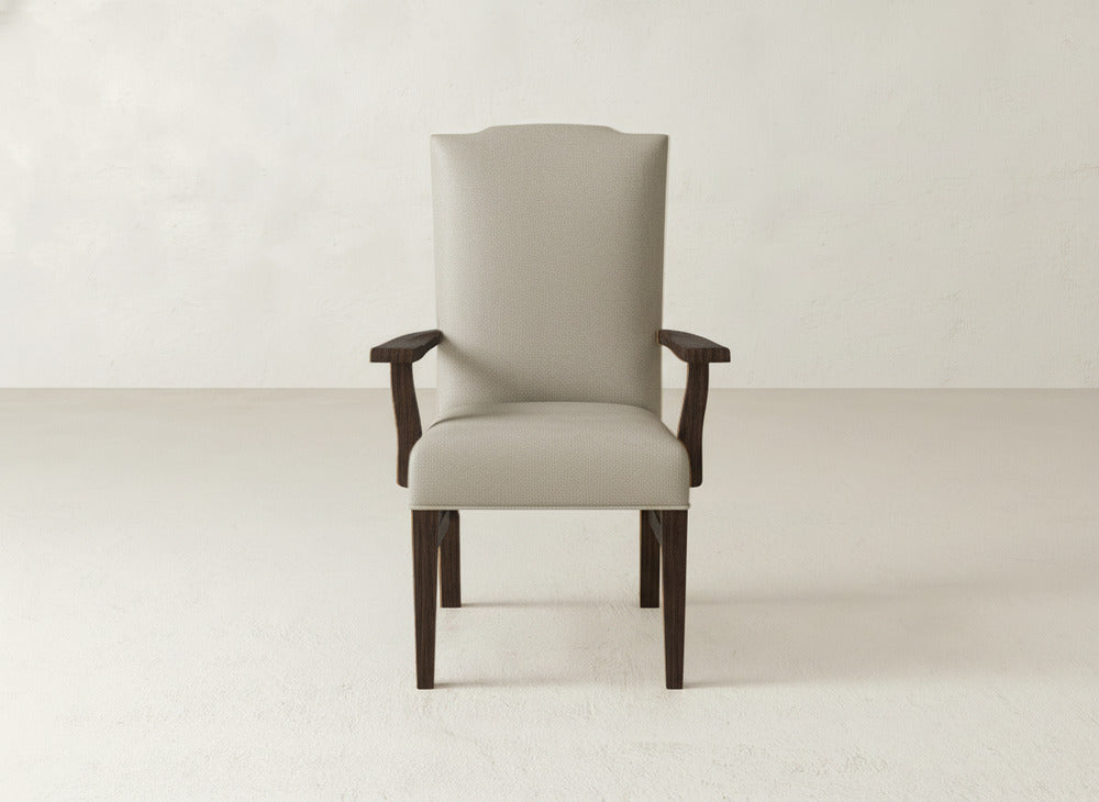 The James+James Hadley Arm Dining Chair is a beige upholstered chair with dark wooden legs and arms, shown facing forward against a plain light wall and floor.
