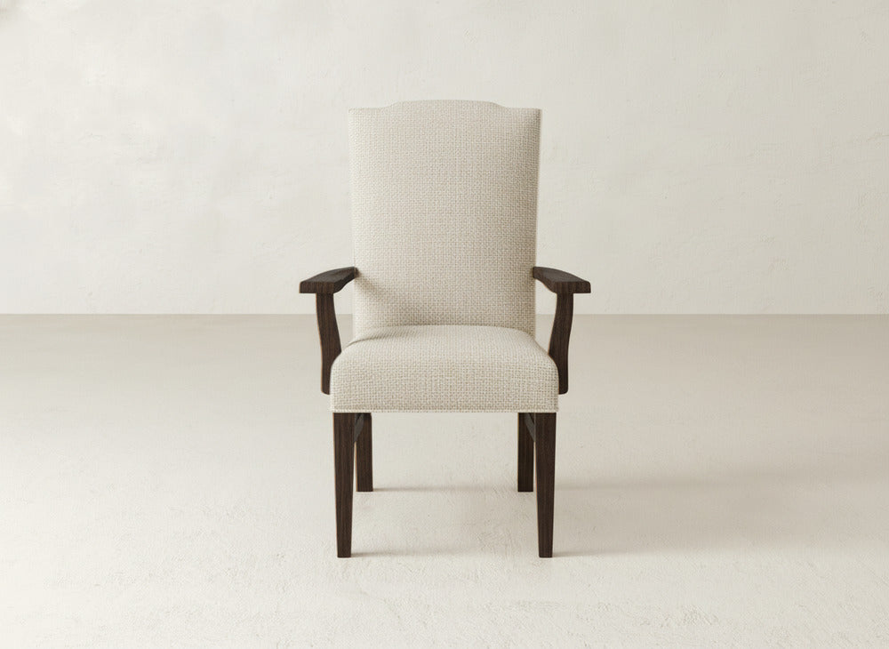 The Hadley Arm Dining Chair by James+James is a beige upholstered piece with a high back, dark wooden legs and armrests, shown against a plain light wall and floor.