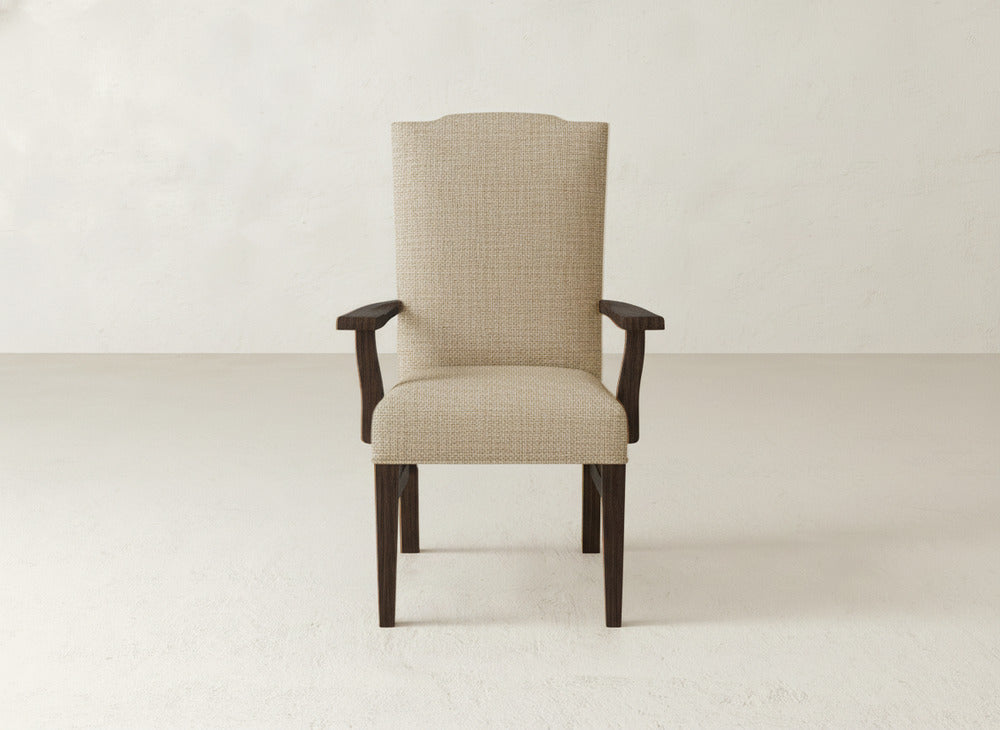 The Hadley Arm Dining Chair by James+James, featuring beige upholstery and dark wooden legs and armrests, is set against a simple light-colored wall and floor backdrop.