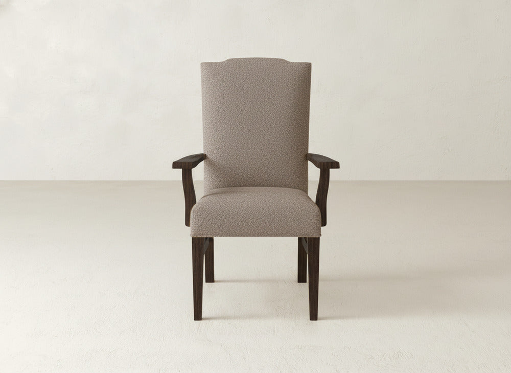 The Hadley Arm Dining Chair by James+James features taupe upholstery, dark wooden legs and arms, a tall curved backrest, and a modern design. It's centered against a plain light wall and floor.