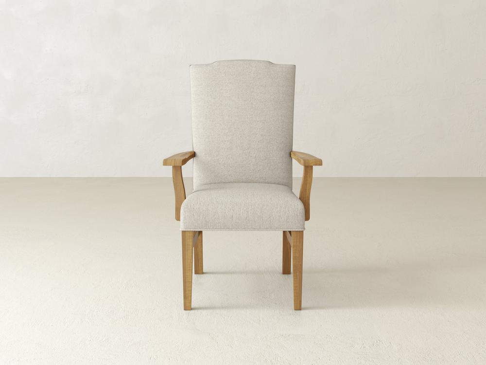The Hadley Arm Dining Chair - Harvest Wheat by James+James is displayed in a minimalistic setting with a light, textured wall and floor, featuring beige upholstery and wooden arms and legs.