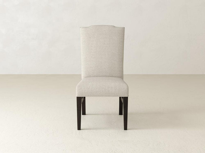 A beige Hadley Side Dining Chair by James+James, featuring a high backrest and dark wooden legs in Charred Ember, set on a light, neutral-colored floor against a plain wall.