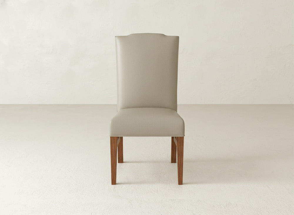 The James+James Hadley Side Dining Chair features a modern high back, light beige upholstery, elegant wooden legs, and subtle tack detailing. It sits on a plain light-colored floor against a neutral wall.