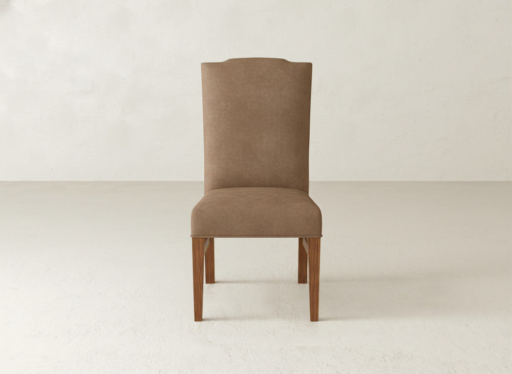 The Hadley Side Dining Chair by James+James features brown upholstery, a high back, and wooden legs. Elegant and versatile, it sits stylishly on a light floor against a plain off-white wall.