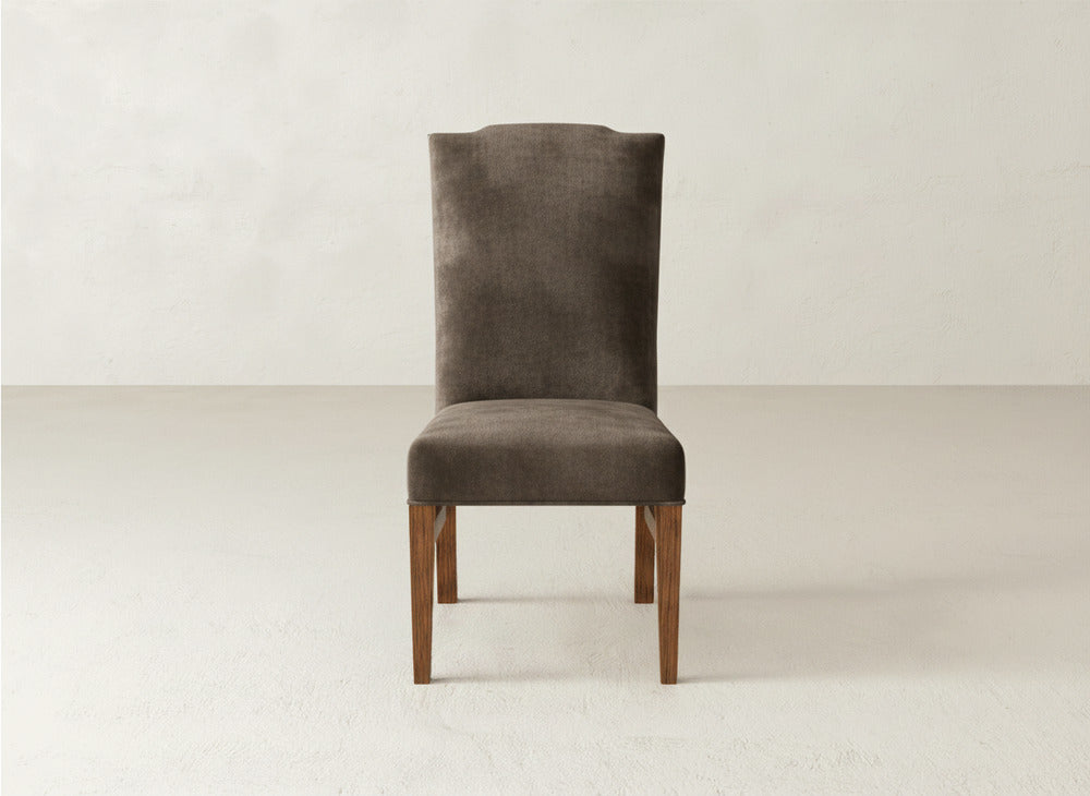 The Hadley Side Dining Chair by James+James, featuring brown upholstery, a high backrest, and wooden legs, is placed on a light-colored floor against a plain, light-colored wall.
