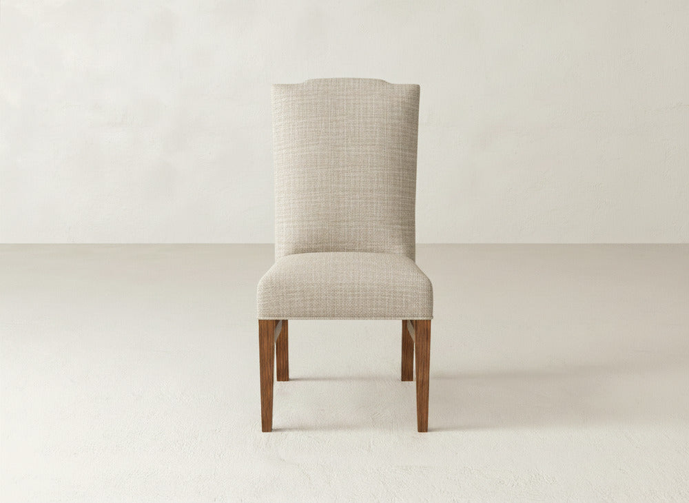 The Hadley Side Dining Chair by James+James, featuring beige upholstery, tack detailing, and wooden legs, is centered against a plain light wall and floor. Its high back highlights a simple, modern design.