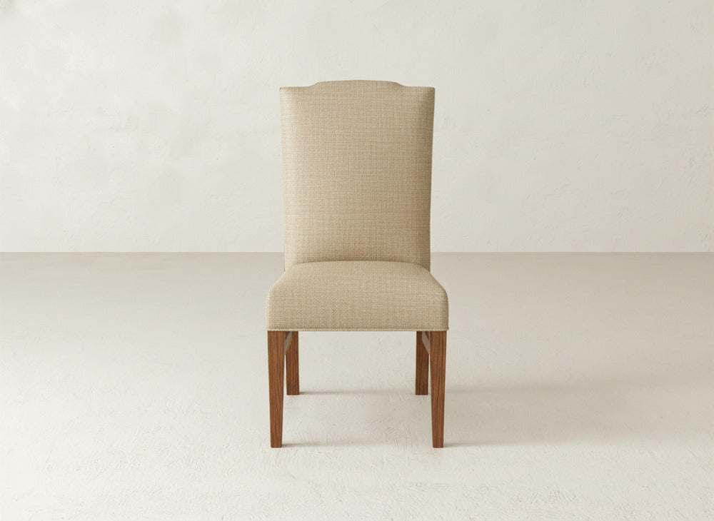 The James+James Hadley Side Dining Chair, featuring beige upholstery, wooden legs, and subtle tack detailing, is centered against a plain white wall and floor background.