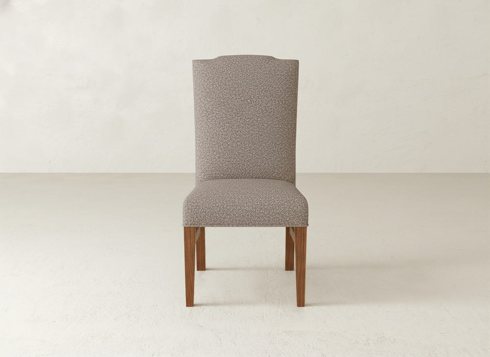 The Hadley Side Dining Chair by James+James, featuring a high back and light patterned upholstery with wooden legs, is centered in a minimal, neutral-toned room with a plain wall and floor.