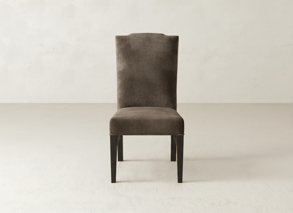 The James+James Hadley Side Dining Chair, with dark brown upholstery, a high backrest, and wooden legs, is positioned on a light floor against a plain off-white wall.