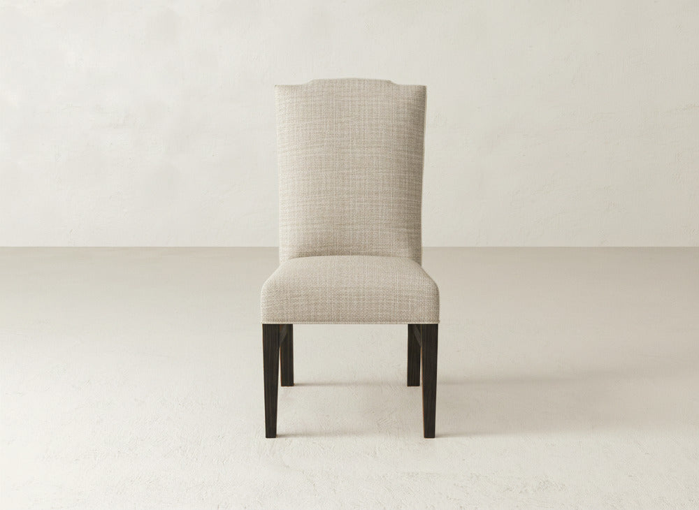 The Hadley Side Dining Chair by James+James features beige upholstery, a high back, and dark wooden legs. This elegant chair is shown on a light floor against an off-white wall.