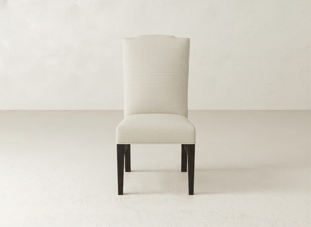 The Hadley Side Dining Chair by James+James features a high back, plush upholstery, and dark wooden legs, set on a light floor against an off-white wall.