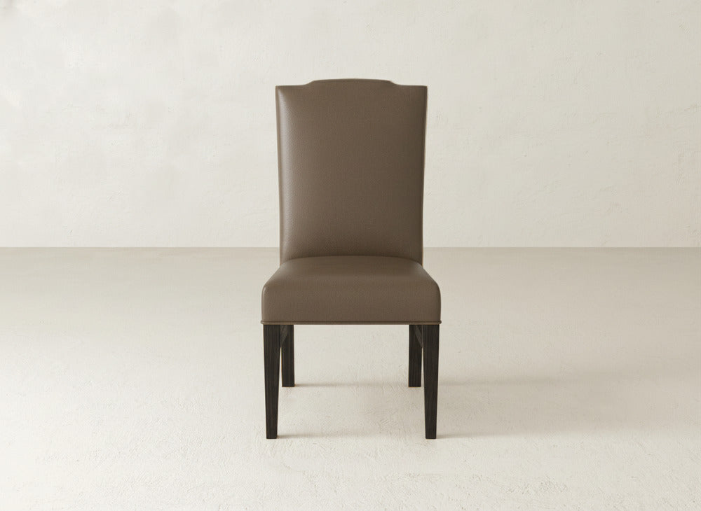 The Hadley Side Dining Chair by James+James features a brown, upholstered high back with elegant tack detailing and dark wooden legs. It’s shown on a light floor against a plain, light wall.