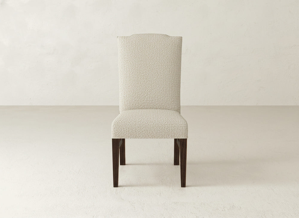 The Hadley Side Dining Chair by James+James features a high, subtly curved upholstered back with refined tack detailing and dark wood legs, all showcased against a plain, light background.