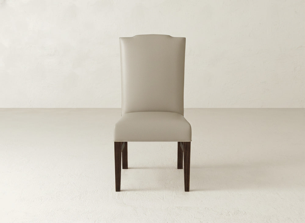 The Hadley Side Dining Chair by James+James, featuring a high back, light beige upholstery, and dark wooden legs, is centered against a plain light-colored wall and floor.