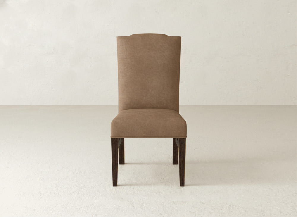 The Hadley Side Dining Chair by James+James features brown upholstery, a tall rectangular backrest, and dark wooden legs. It is shown on a light floor against a plain off-white wall.