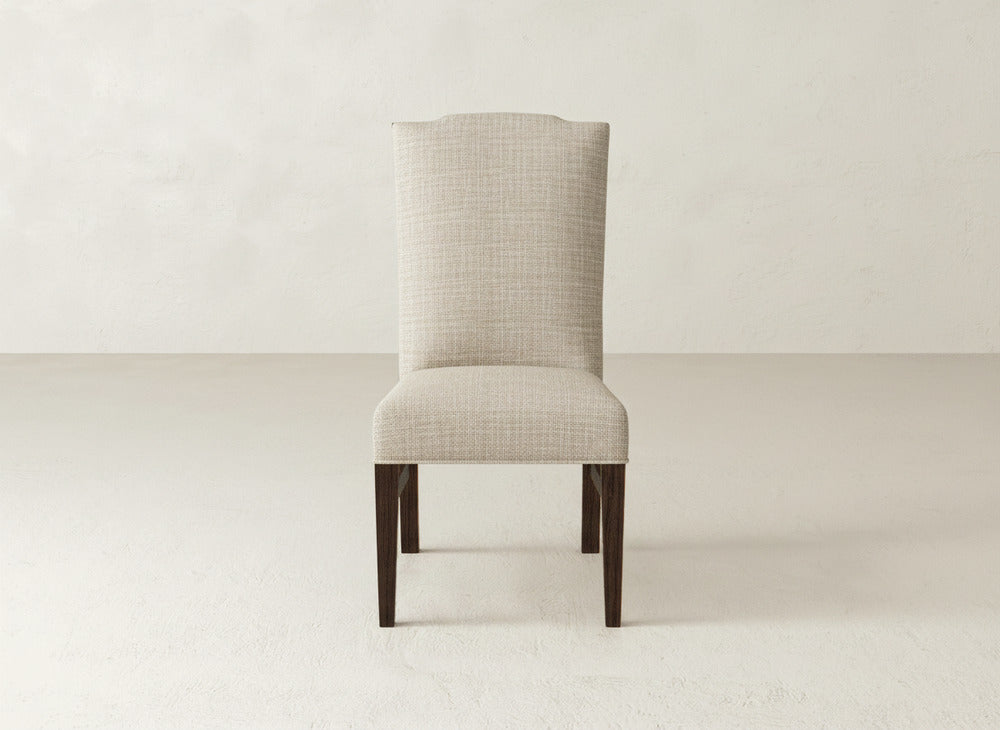 The Hadley Side Dining Chair by James+James features a neutral-toned, upholstered high back and dark wood legs, bringing elegance to any dining space when placed against light walls and flooring.