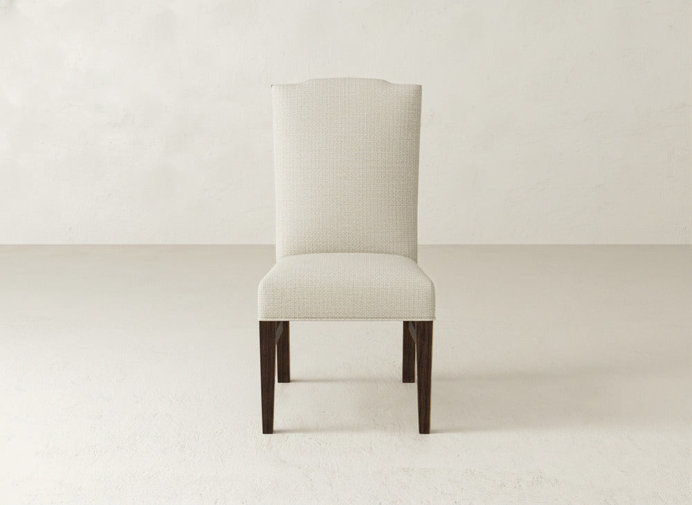 The Hadley Side Dining Chair by James+James features a beige high back with subtle tack detailing, a tall, gently curved backrest, and dark wooden legs, set on a light floor against a plain light wall.