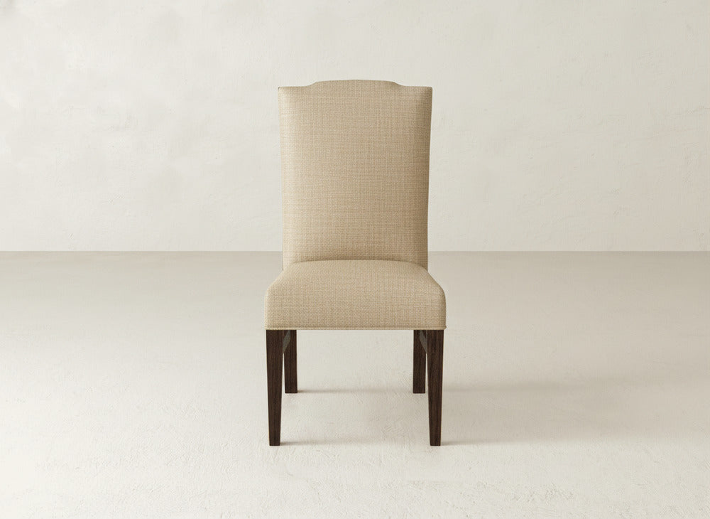 The Hadley Side Dining Chair by James+James features beige upholstery, a tall slightly curved high back, and dark wooden legs, shown on a light floor against an off-white wall.