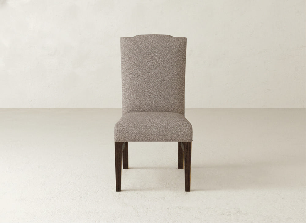 The James+James Hadley Side Dining Chair features a tall upholstered back with subtle patterned fabric, tack detailing along the edges, and dark wooden legs, shown in a minimal, light-hued room.