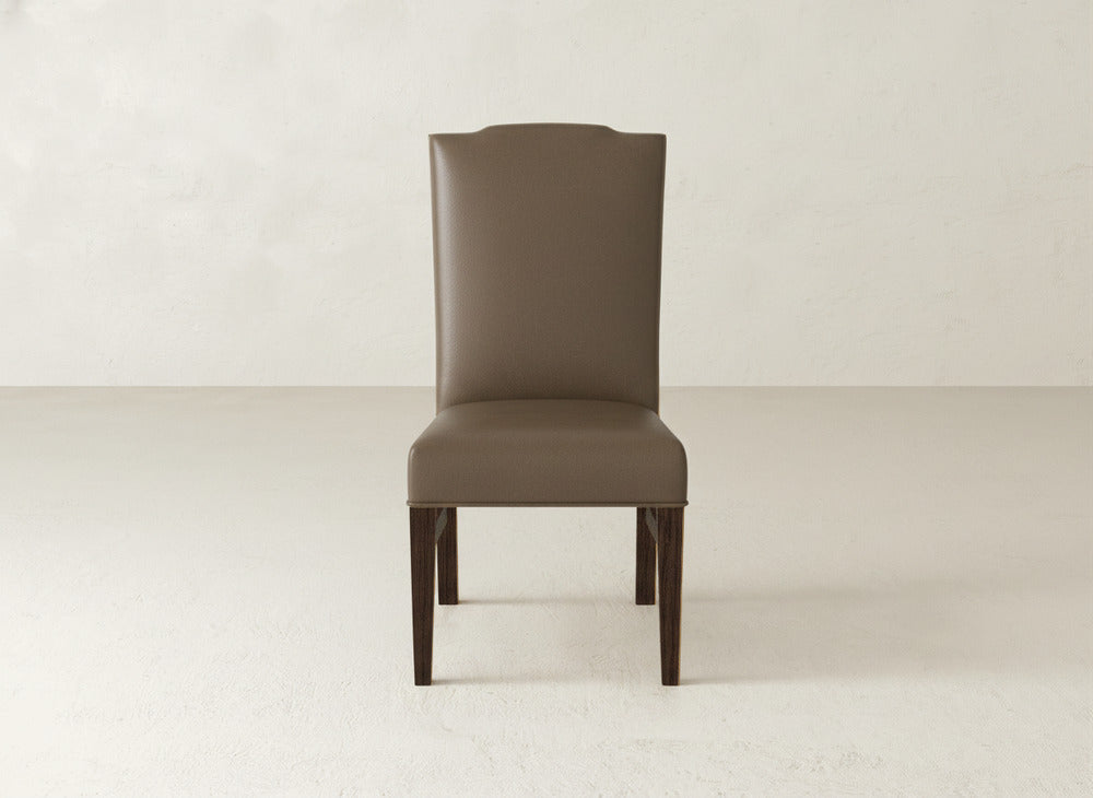 The Hadley Side Dining Chair by James+James, with brown upholstery, tack detailing, a high back, and dark wooden legs, is placed on a light floor against a plain light wall.
