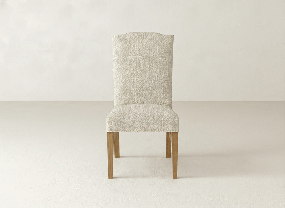 The Hadley Side Dining Chair by James+James, featuring light textured upholstery and wooden legs, sits in a minimalist room with neutral walls and flooring.
