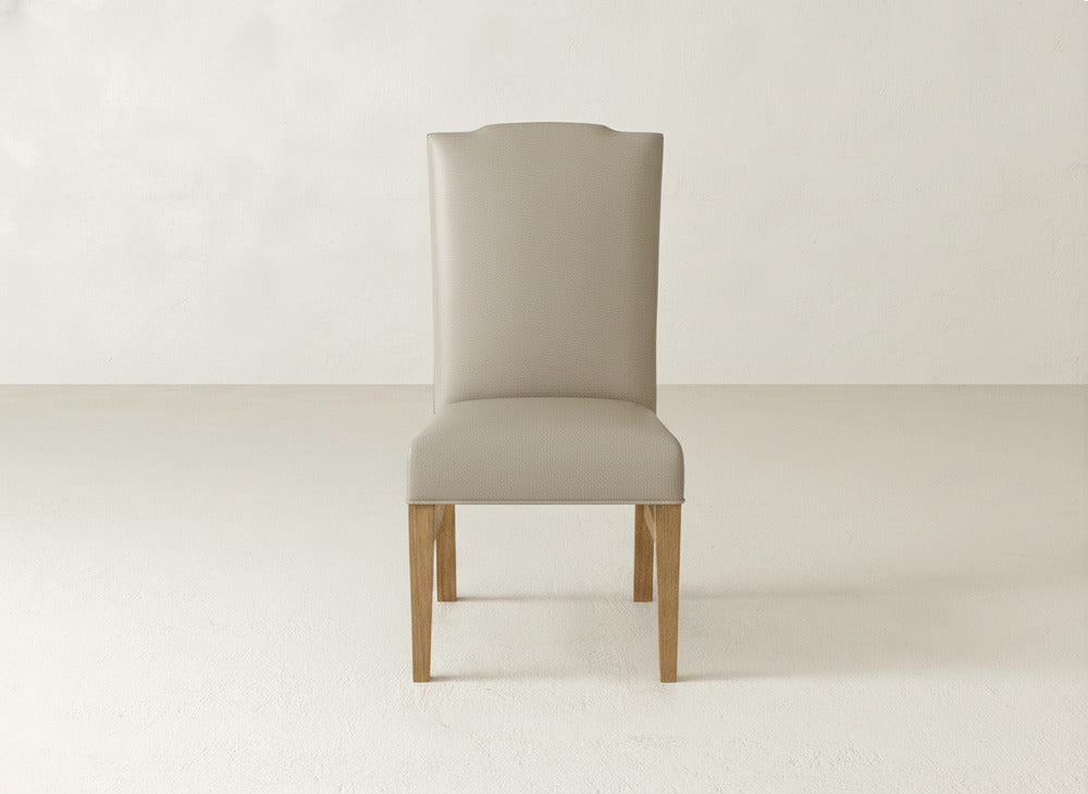 The Hadley Side Dining Chair by James+James, featuring a beige high back and wooden legs, is centered against a plain, light-toned wall and floor, highlighting its elegant silhouette.