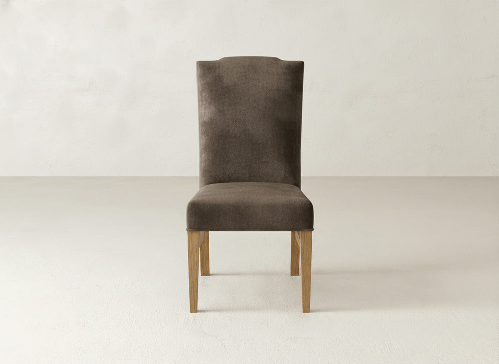 The Hadley Side Dining Chair by James+James is a brown high-back chair with elegant upholstery, subtle tack accents, and light wooden legs, displayed against a plain light background.
