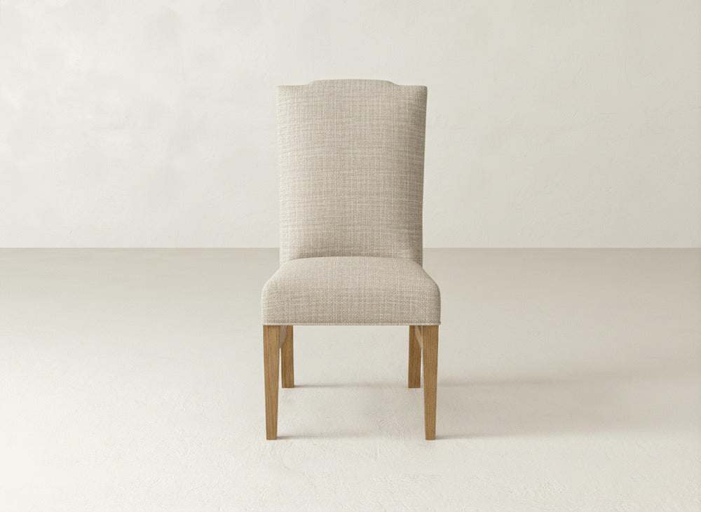 The Hadley Side Dining Chair by James+James features a beige upholstered seat, high curved backrest with subtle tack details, and wooden legs, shown on a light floor against an off-white wall.