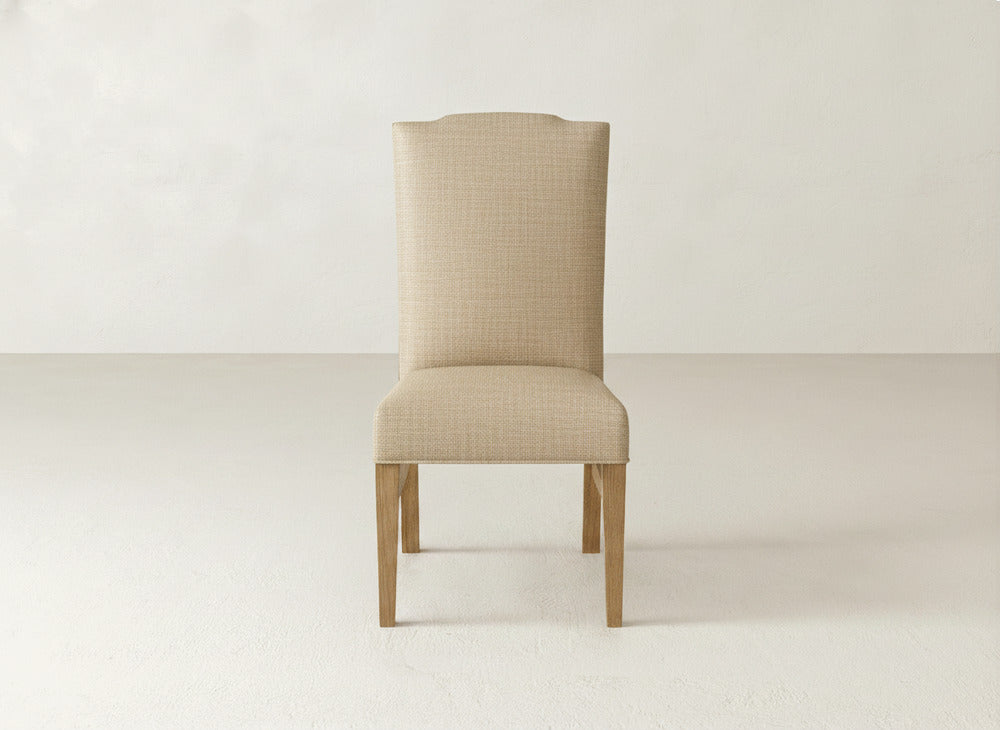 The Hadley Side Dining Chair by James+James features beige upholstery with tack detailing, a high backrest, and wooden legs, shown against a simple light-colored background.