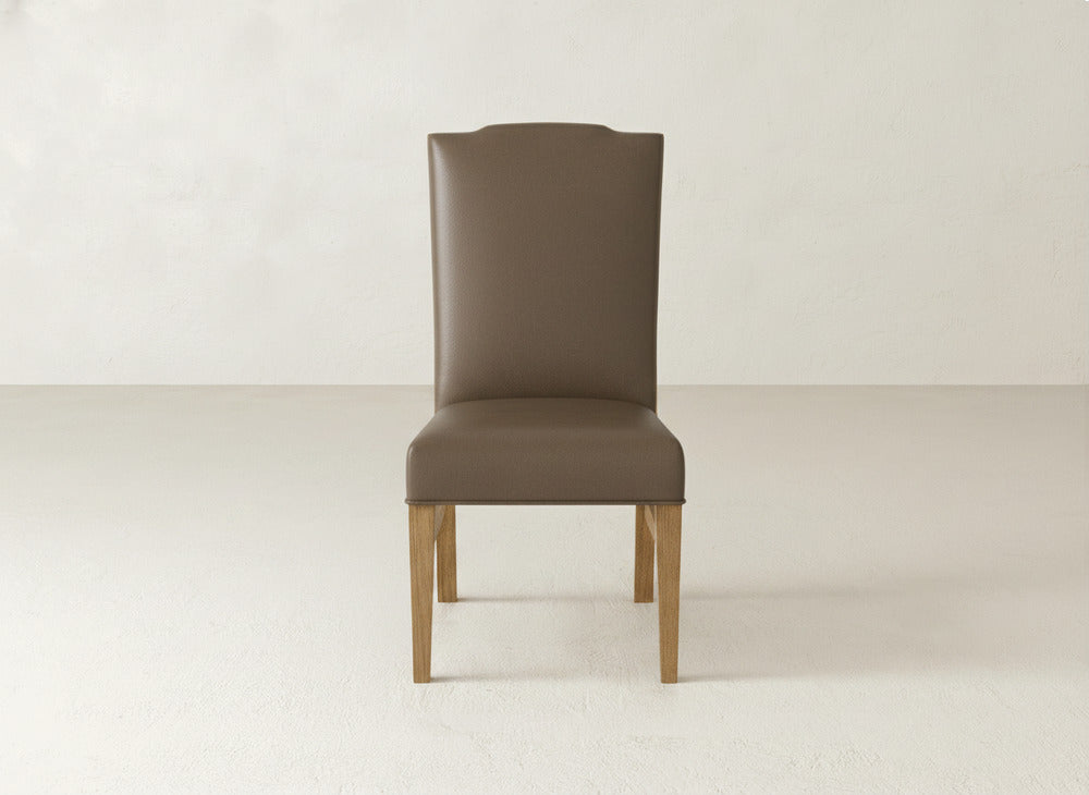 The Hadley Side Dining Chair by James+James features a high back, brown fabric upholstery, elegant tack detailing, and wooden legs. It sits on a light floor with a plain off-white background.