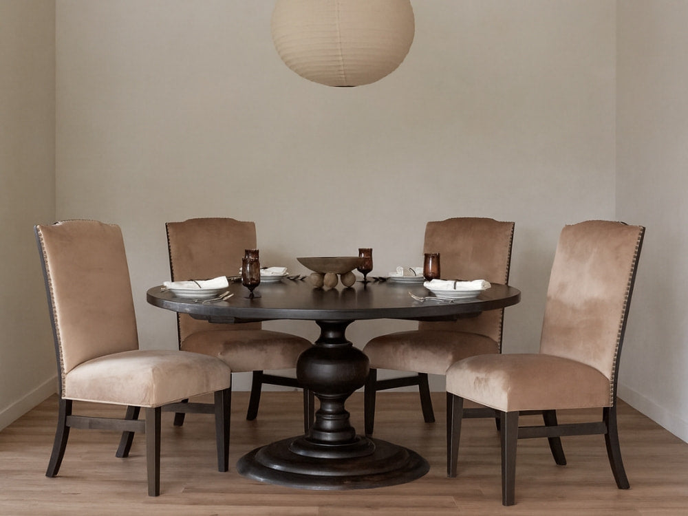 A round dark wood dining table with four James+James chairs in Latte velvet sits on a light wood floor. The table is set with napkins, glasses, and a bowl, and a large round paper pendant hangs above.