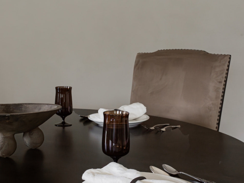 A dark wood dining table is set with white napkins, silverware, plates, and two dark glasses. Behind the table sits a James+James Latte velvet chair, featuring a caramel tone and soft hand, against a plain light wall.