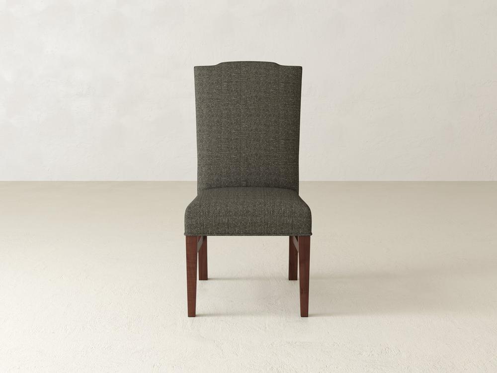 The Hadley Side Dining Chair - Tuscany by James+James, featuring a high back with dark gray upholstery and wooden legs, is positioned on a beige floor against a plain light-colored wall.