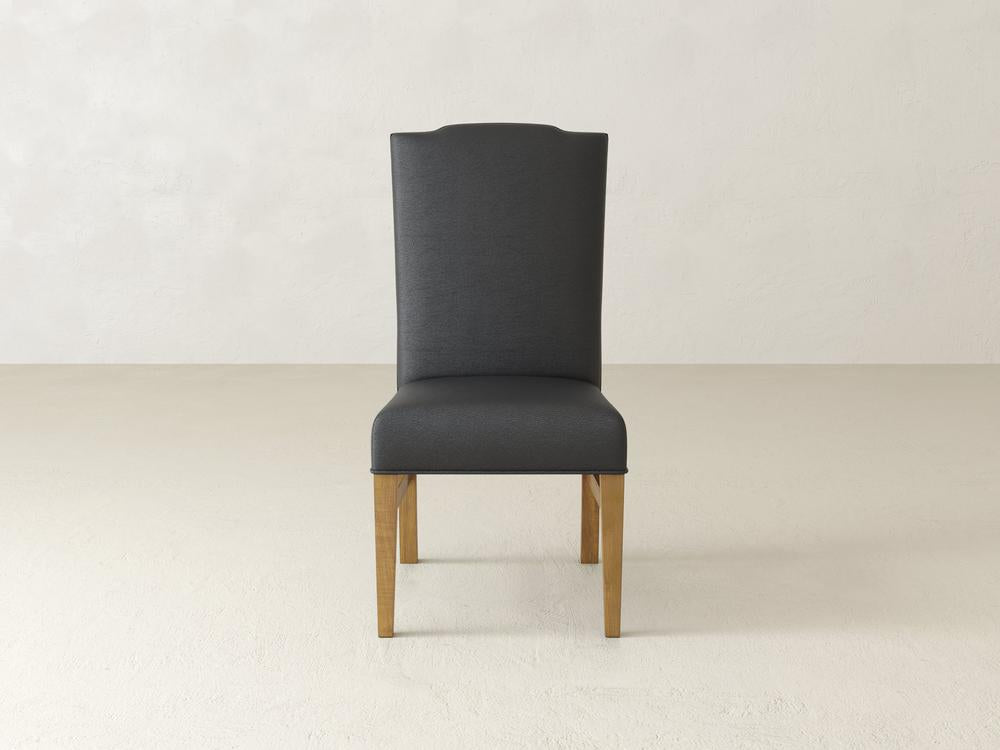 A modern high-backed chair with dark upholstery and wooden legs stands on a light, textured floor against a plain, light-colored background.